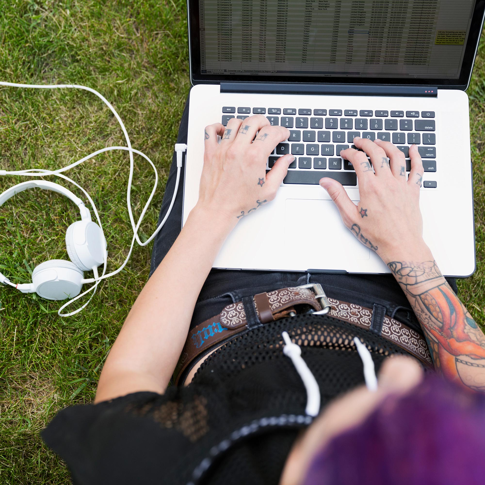 Ung person med tatueringar sitter ute i gräset med en laptop i knät